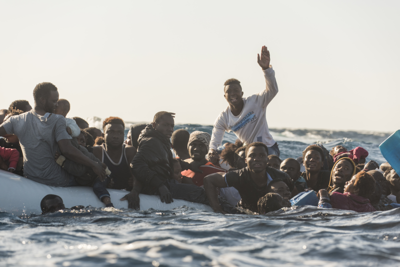 Ein sinkendes Boot voller ermüdeter, verzweifelter Menschen wurde entdeckt. Eine Person winkt den Rettern zu.