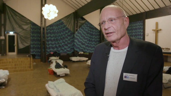 Ein Mann mit Brille und Namensschild steht in einem Raum voller Feldbetten. Im Hintergrund sind ein Kreuz und eine Tür zu sehen.